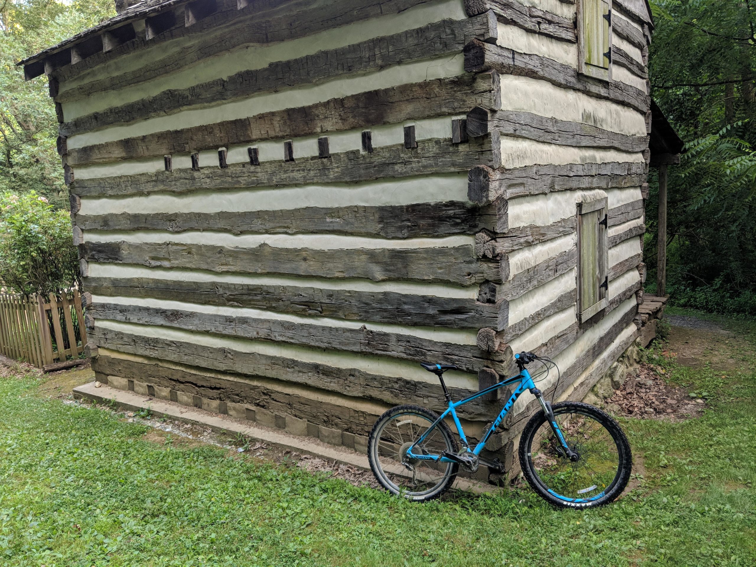 A blue mountain bike is resting against the side of a rustic log cabin, which features alternating light and dark wooden logs and small windows with wooden shutters. The cabin is surrounded by lush greenery and a wooden fence, with a pathway visible in the background. Hashawha mountain bike trail.