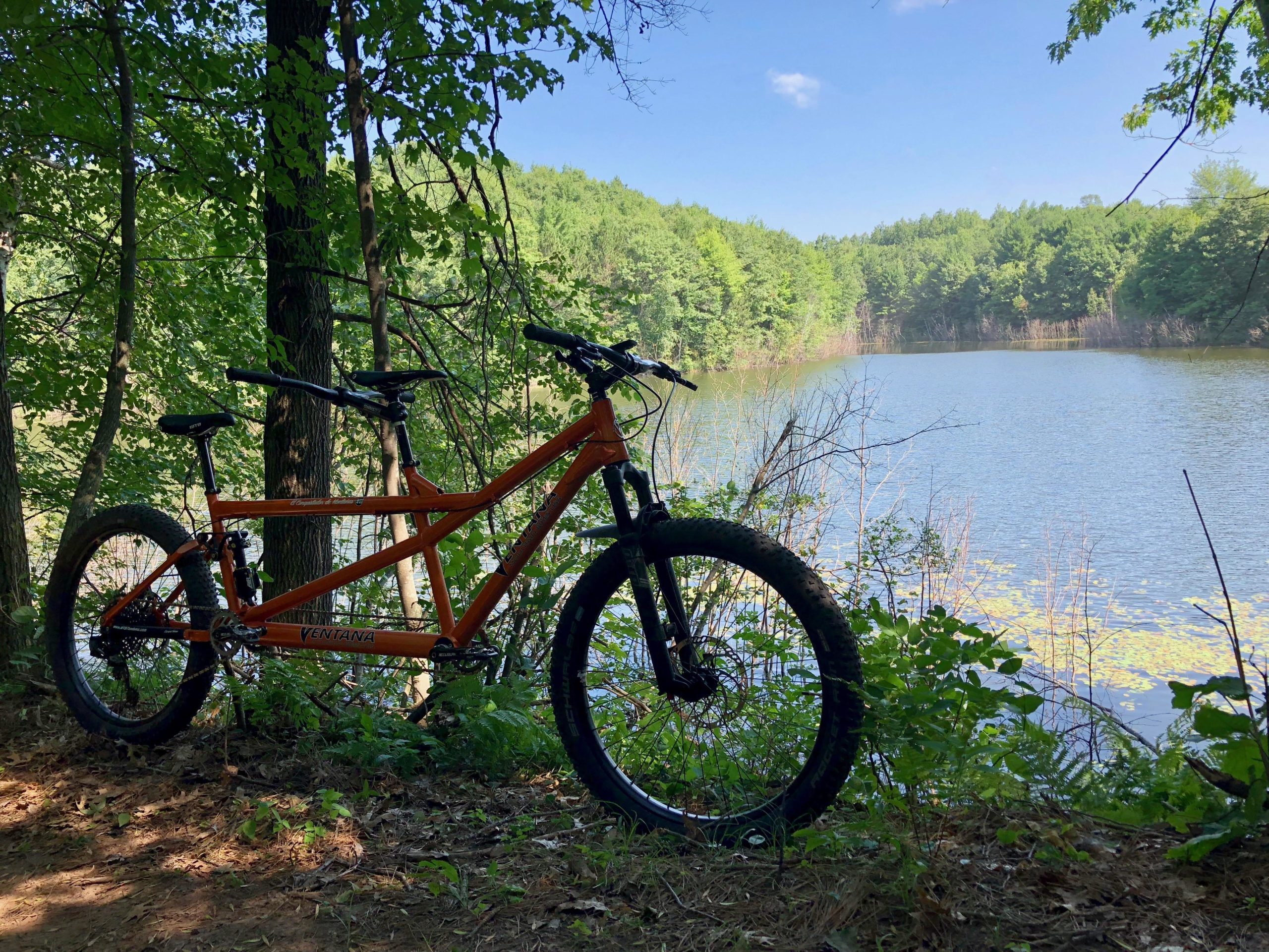A vibrant orange tandem mountain bike rests beside a serene lake, surrounded by lush green trees. The calm water reflects the blue sky, creating a peaceful outdoor scene. Hickory Ridge mountain bike trail.