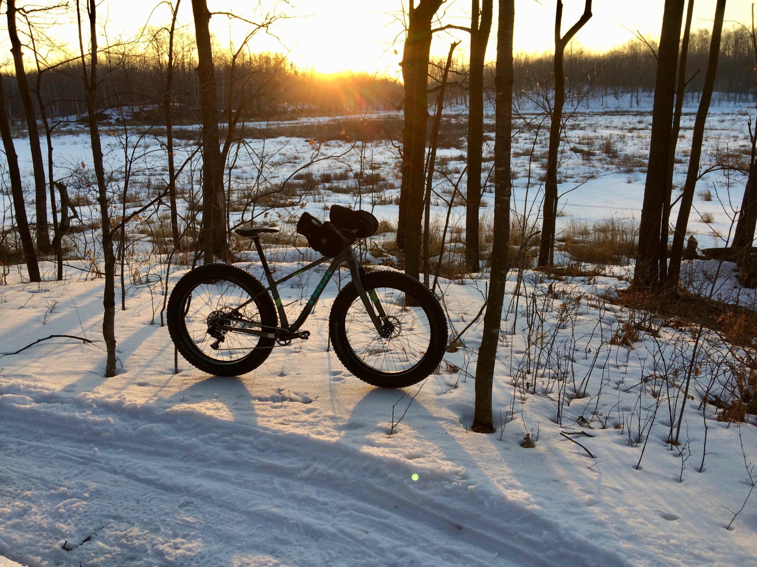 A fat tire bicycle parked on a snowy trail surrounded by trees, with a golden sunset casting warm light across the scene. The landscape features a frozen, grassy area in the background, highlighting a serene winter atmosphere. Hickory Ridge mountain bike trail.