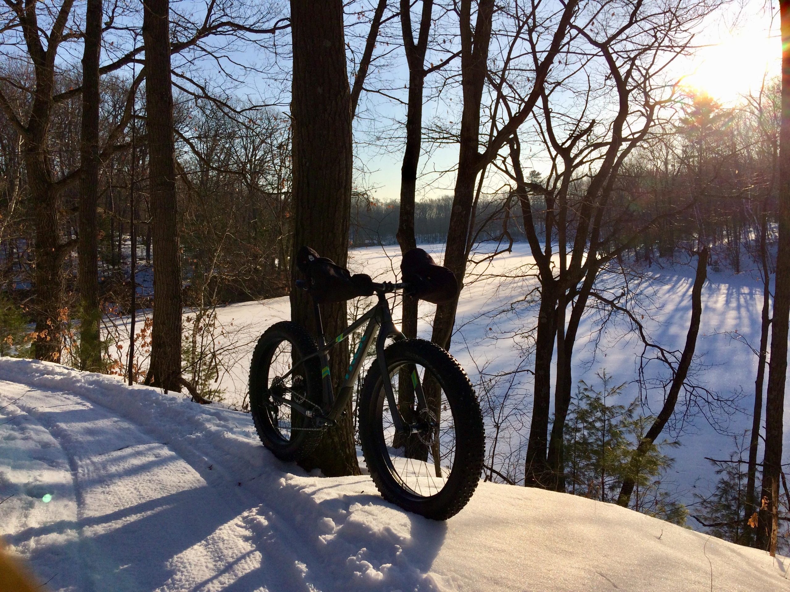 A fat bike rests on a snowy trail surrounded by trees, with a frozen lake visible in the background. The scene is illuminated by bright sunlight, casting long shadows on the snow. Hickory Ridge mountain bike trail.