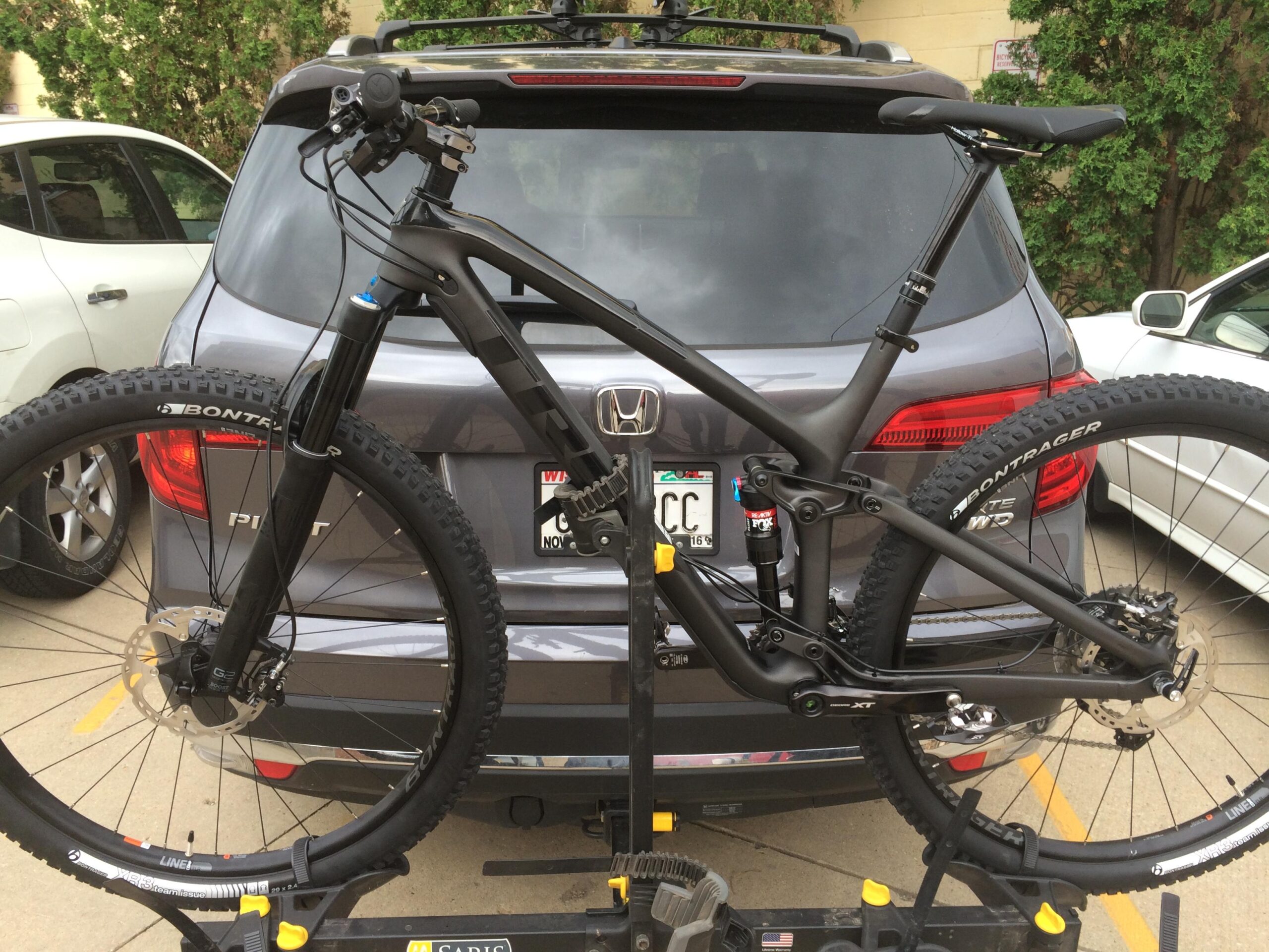 Trek Fuel EX 9.8: A black mountain bike is mounted on a bike rack attached to the back of a gray Honda SUV. The bike features thick tires and a front suspension fork, showcasing its off-road capabilities. The SUV, parked in a lot, has visible taillights and a license plate, with a second vehicle seen in the background.