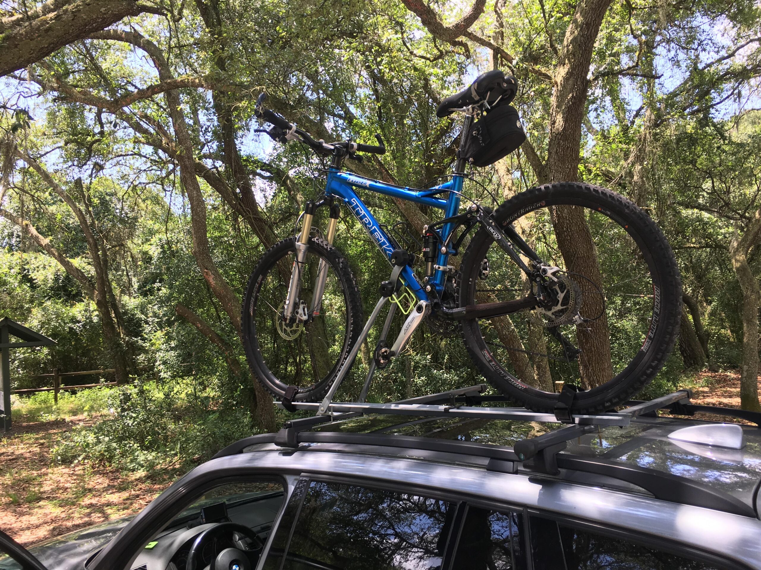 Trek Fuel EX 9: A blue mountain bike mounted on the roof rack of a vehicle, surrounded by trees in a wooded area.