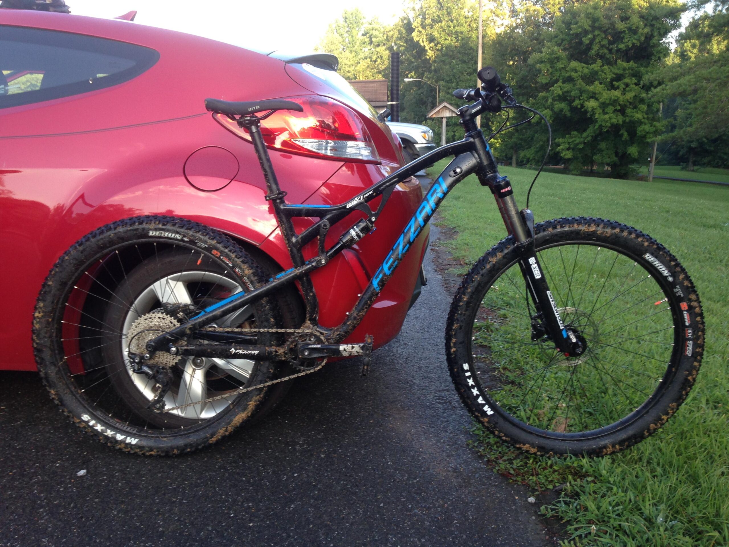 Fezzari Wiki Peak: A black mountain bike with blue accents is parked next to a red car on a paved surface. The bike features muddy tires, indicating recent use on a trail, while the background includes green grass and trees. The setting suggests an outdoor adventure location.