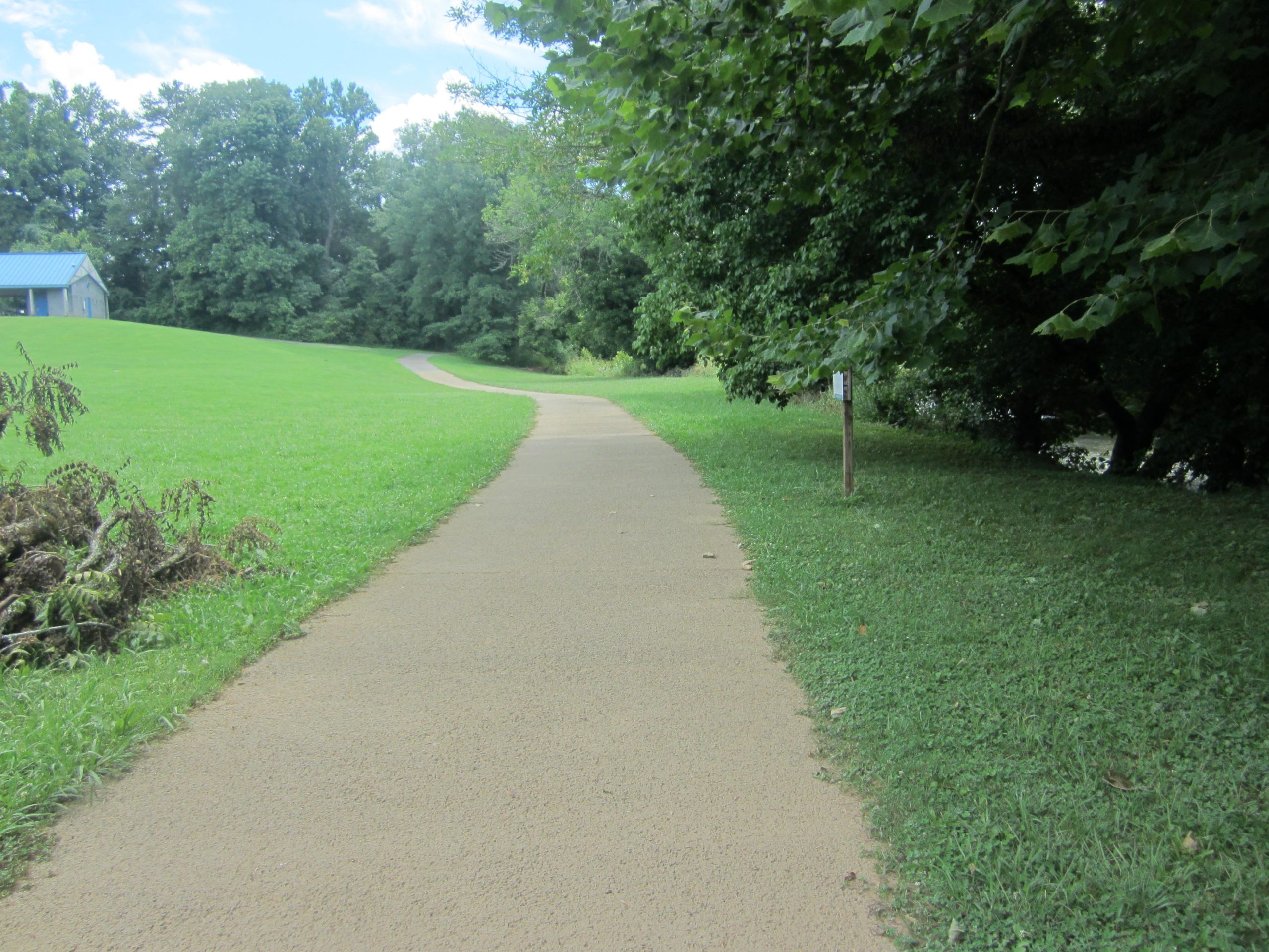A paved pathway meanders through a green landscape, flanked by grass and trees. A small building with a blue roof is visible in the distance to the left. The scene conveys a serene outdoor setting under a partially cloudy sky. Athens Regional Park Trail mountain bike trail.