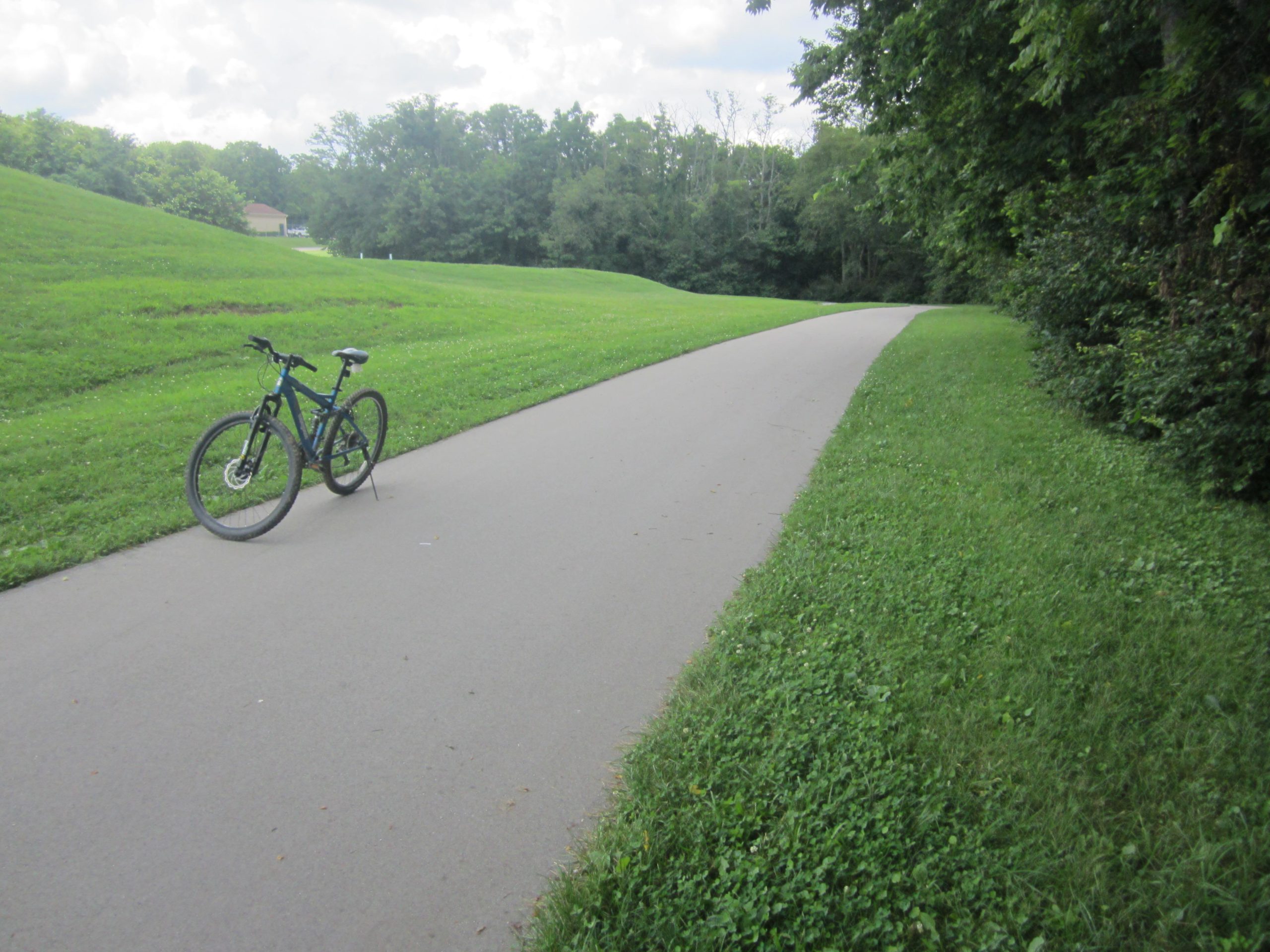 A mountain bike resting on a paved pathway beside a lush, grassy area, surrounded by trees under a partly cloudy sky. Veterans Park mountain bike trail.