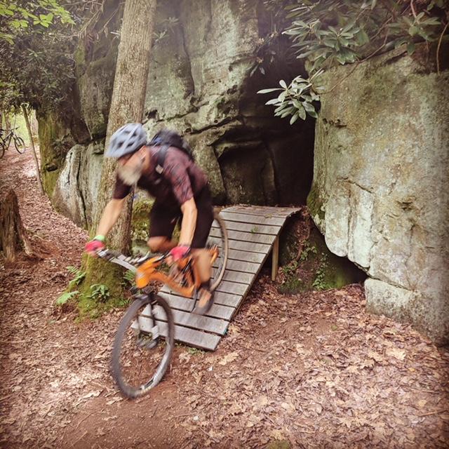 A mountain biker with a helmet rides over a wooden ramp on a trail surrounded by lush greenery and large rock formations. The ground is covered in brown leaves, and trees are visible in the background. Big Bear Lake Trail Center mountain bike trail.