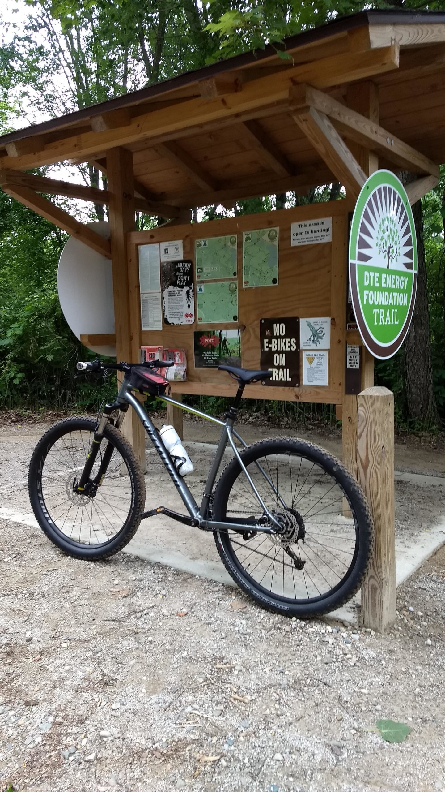 A mountain bike parked next to a wooden trailhead sign for the DTE Energy Foundation Trail, featuring maps and trail information, surrounded by trees. DTE Energy Foundation Trail mountain bike trail.