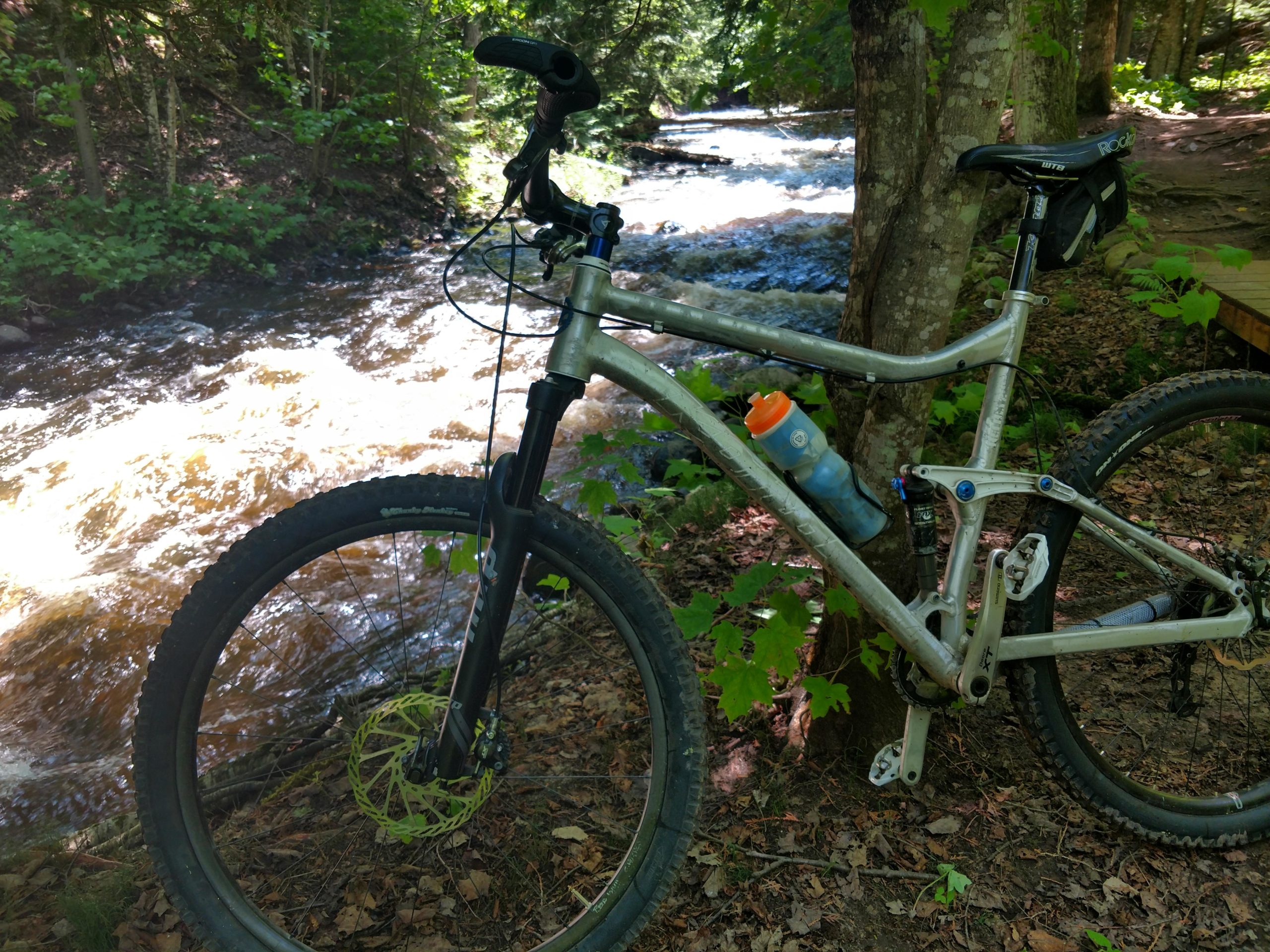 A mountain bike parked beside a flowing creek, surrounded by lush greenery and trees. The bike features a water bottle mounted on the frame, and the creek has a brownish tint, suggesting recent rainfall. Noquemanon Trails Network: South Marquette Trails mountain bike trail.