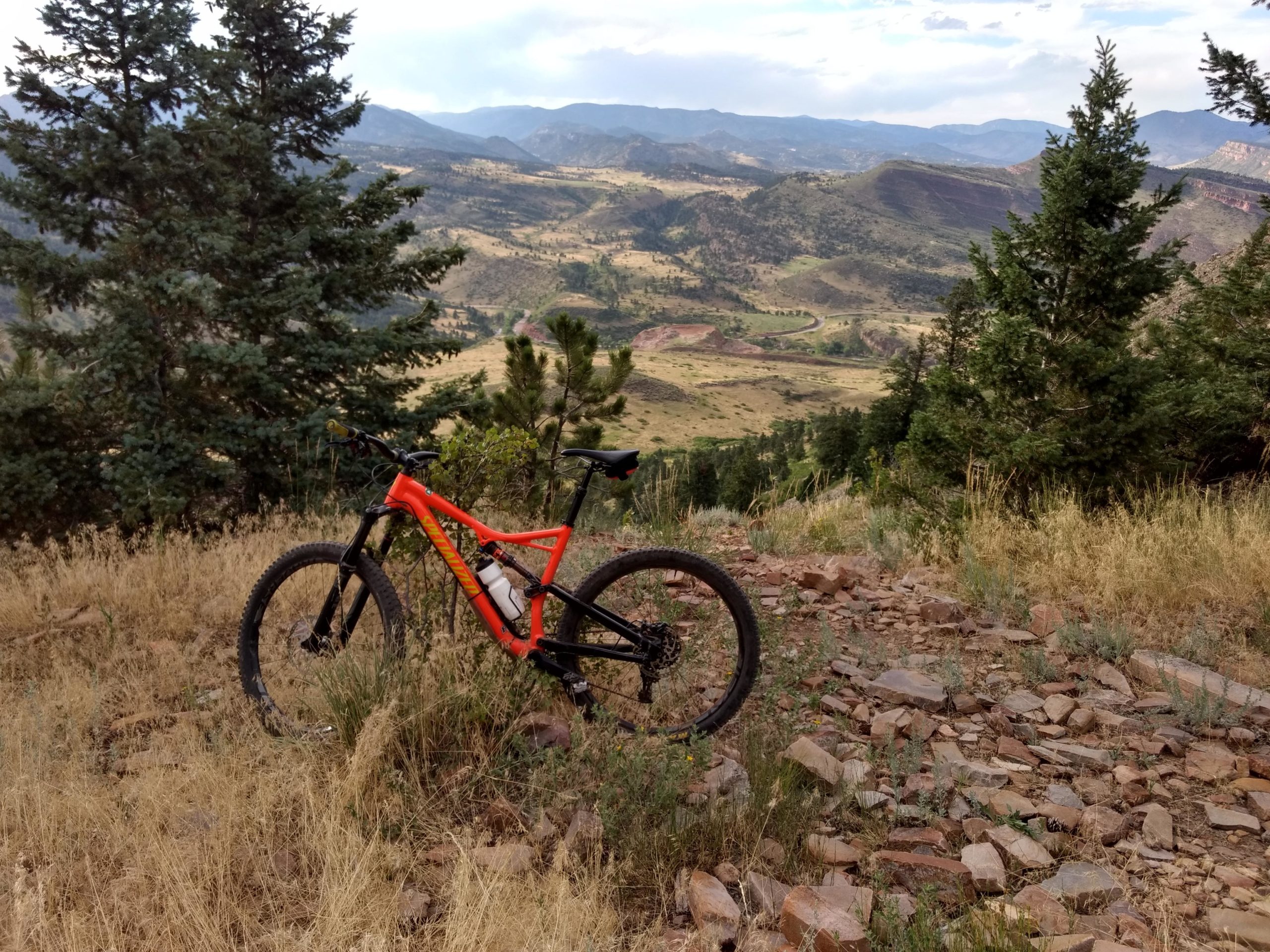 A bright orange mountain bike is positioned on a rocky trail overlooking a scenic landscape of rolling hills and distant mountains, surrounded by pine trees and tall grass. The sky is partly cloudy, hinting at a beautiful outdoor adventure. Heil Valley Ranch mountain bike trail.