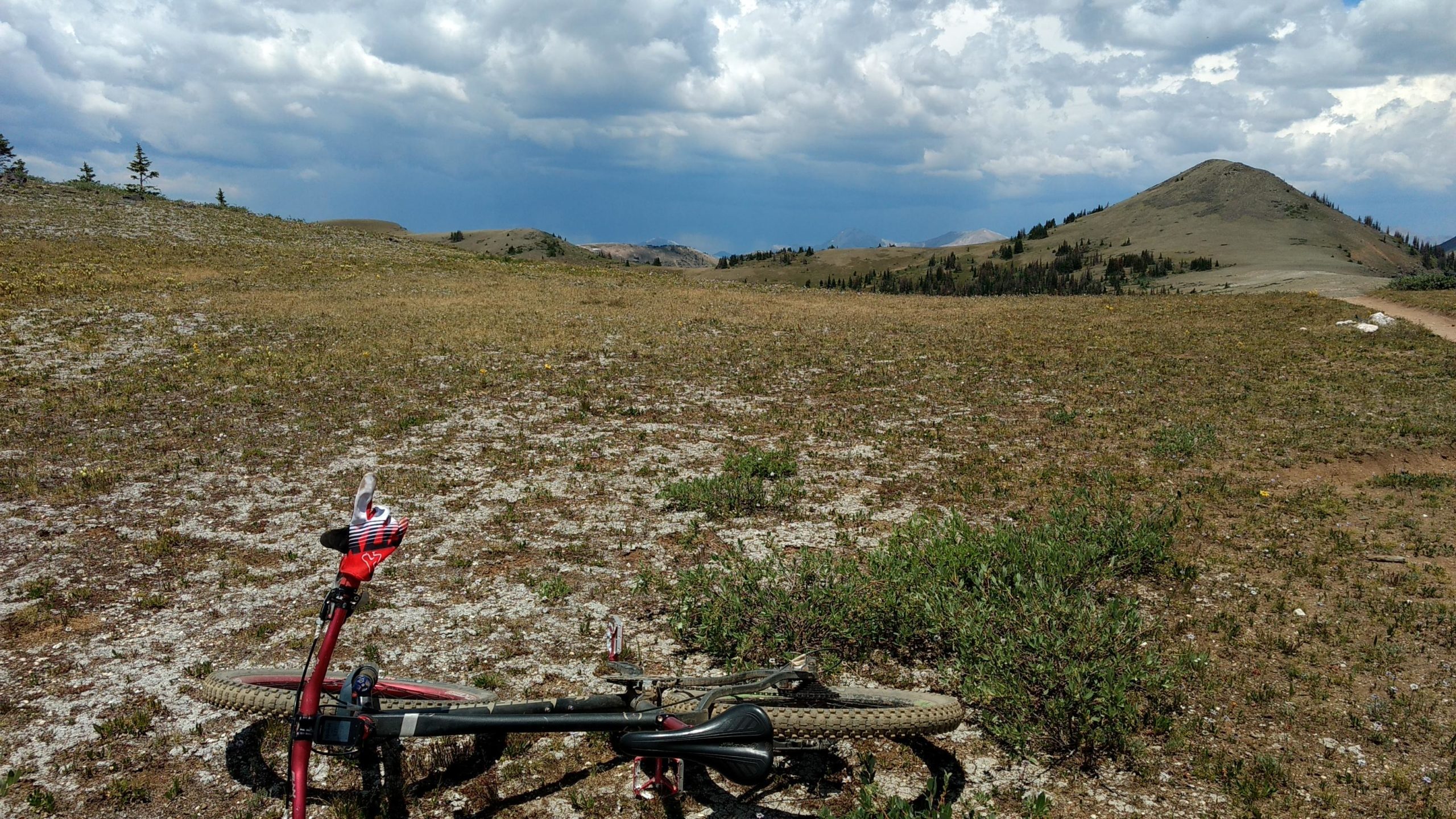 A mountain bike resting on a rocky, grassy terrain with rolling hills and a cloudy sky in the background. Monarch Crest Trail mountain bike trail.