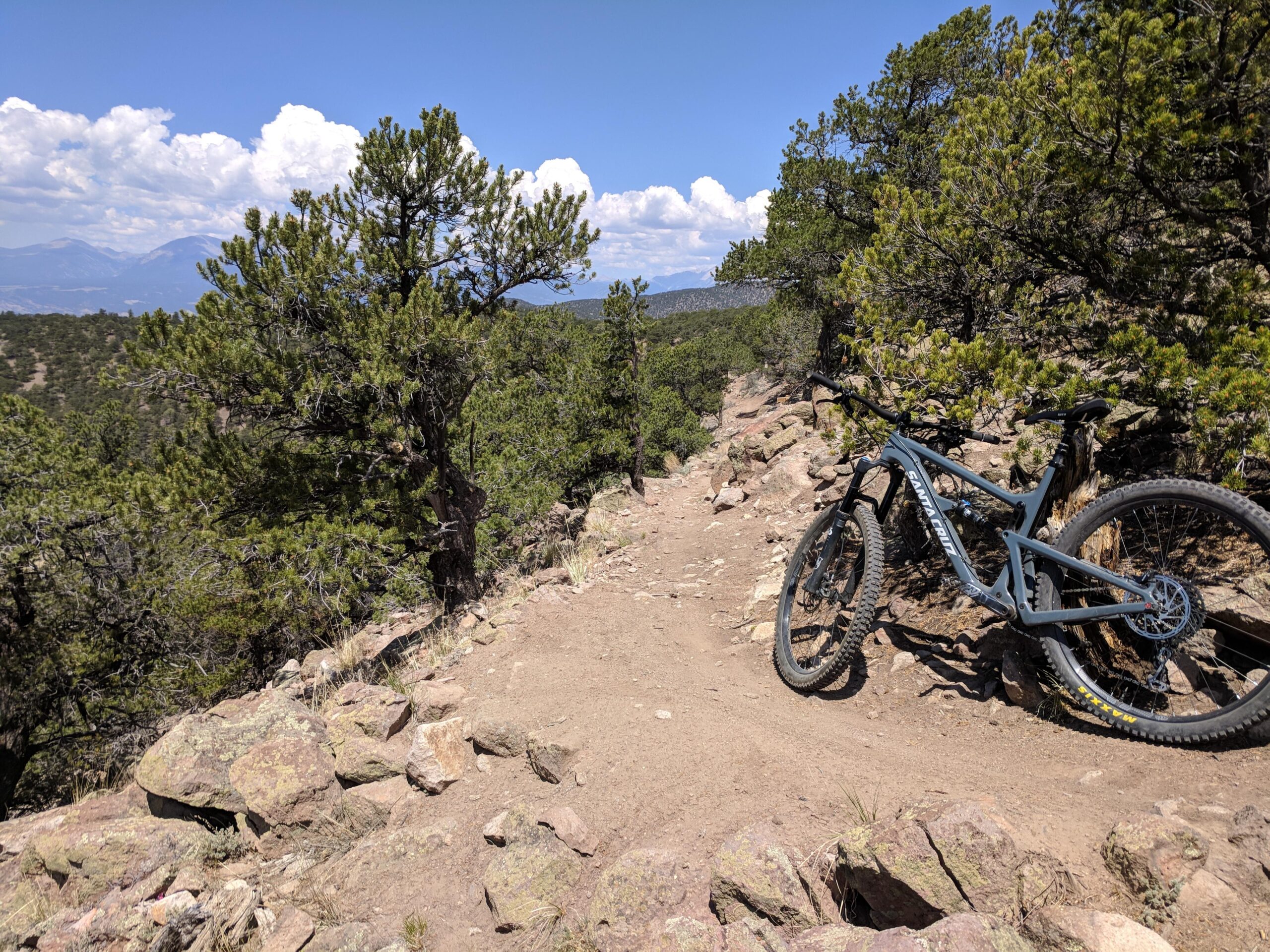 Santa Cruz Hightower LT: A mountain bike resting on a rocky trail surrounded by trees, with distant mountains and a blue sky filled with clouds in the background.