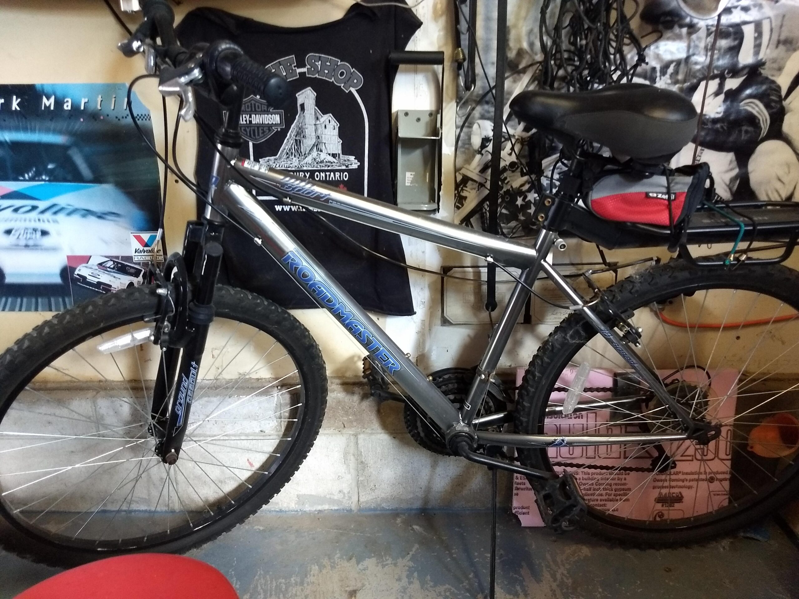 Trek Marlin 5: A silver mountain bike with thick tires is leaning against a wall in a garage. The bike features a front suspension fork and a red saddle bag attached to the back. Behind the bike, there are various posters and tools hanging on the wall, contributing to the garage's cluttered environment. The floor is made of concrete, and there is a red chair partially visible in the foreground.
