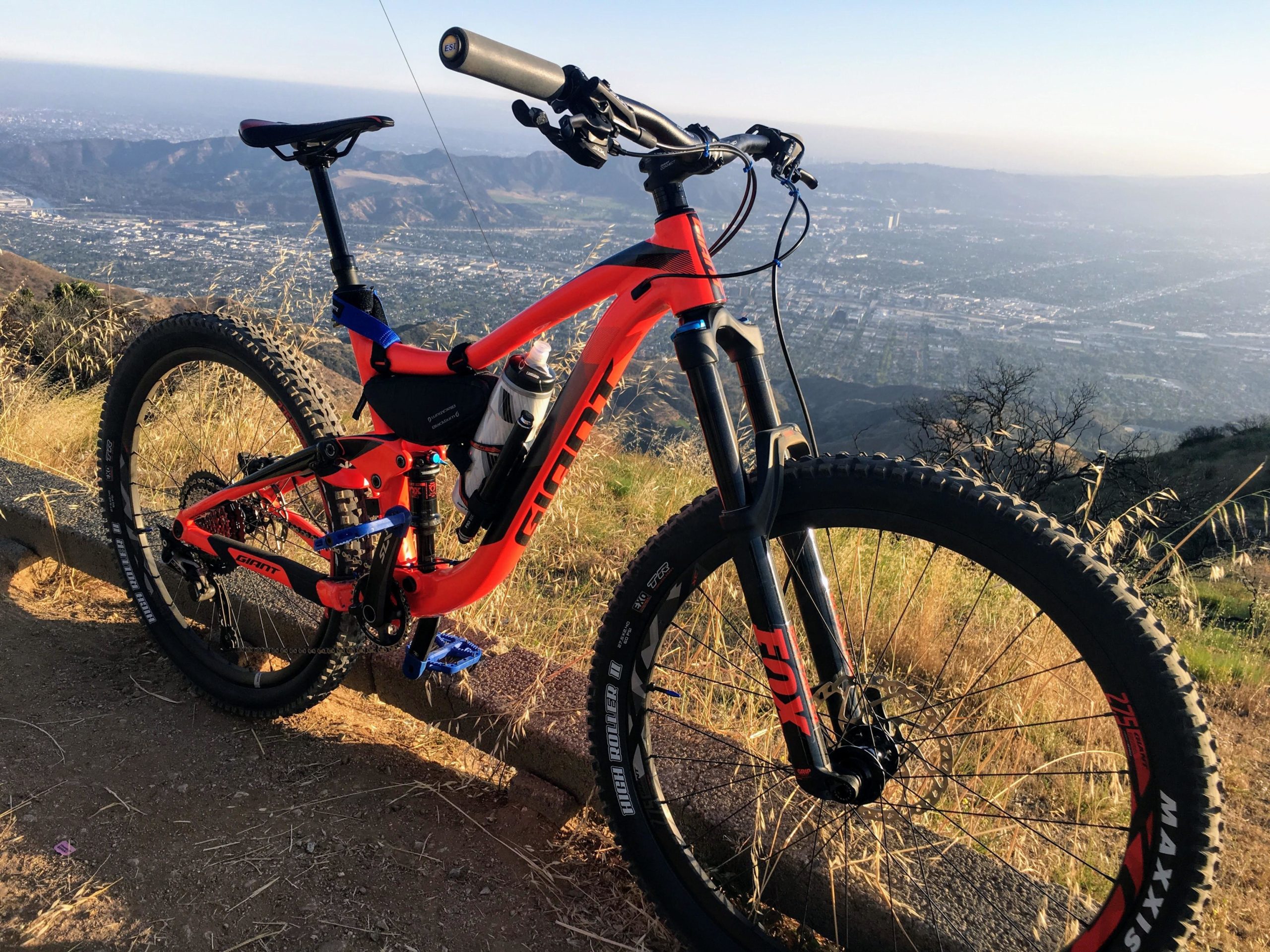 A brightly colored orange mountain bike parked on a rocky trail, overlooking a scenic valley and distant mountains. The bike features a water bottle holder and is set against a backdrop of grass and hills, with a panoramic view of the city below. La Tuna Canyon mountain bike trail.