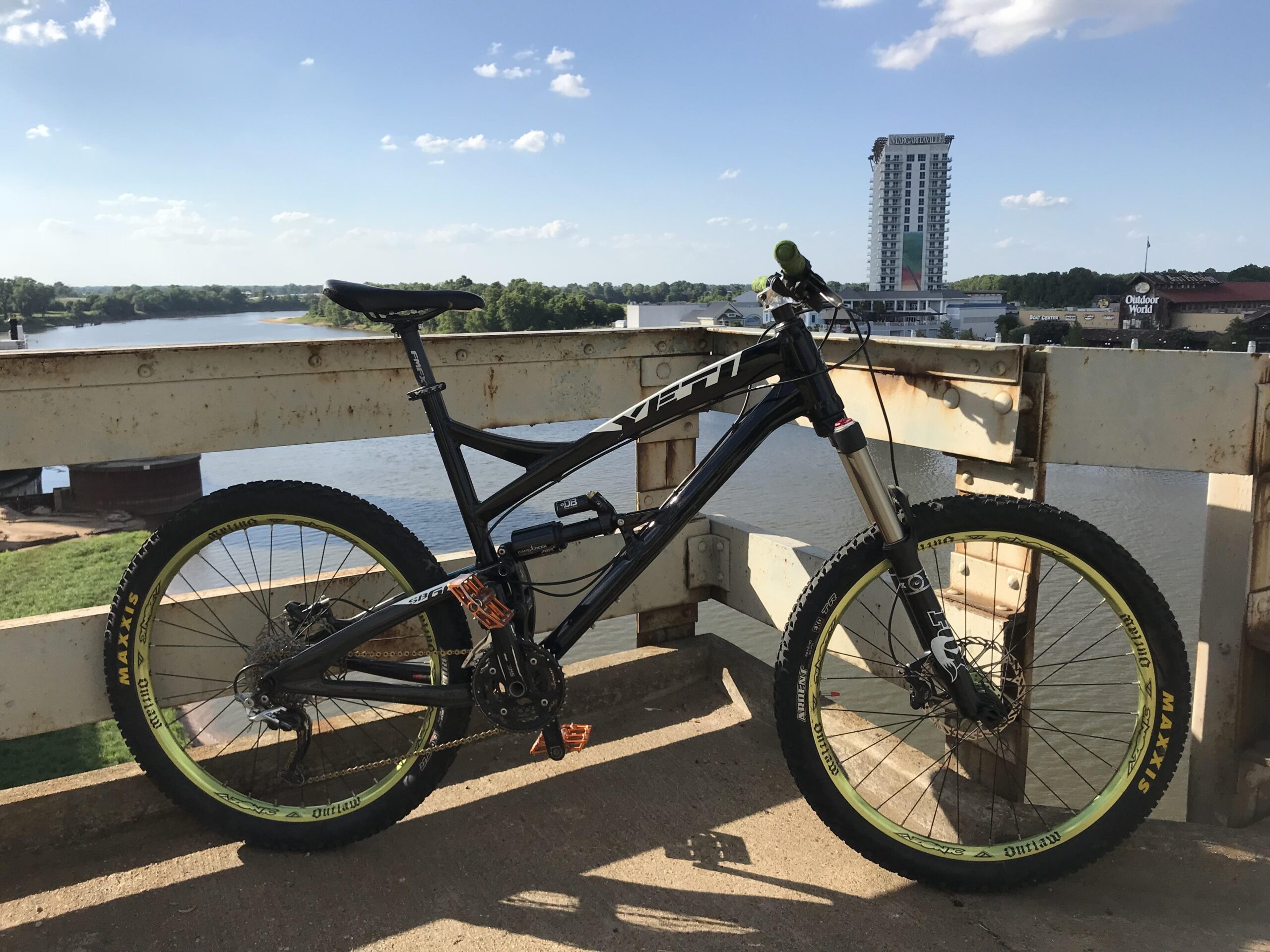 Yeti SB66: Mountain bike resting on a bridge railing with a river and trees in the background, under a clear blue sky with scattered clouds. A tall building is visible in the distance, along with some commercial structures near the water.
