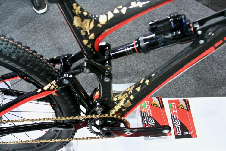Close-up view of a modern mountain bike frame featuring a combination of black and red paint, with decorative gold accents. The image showcases the bike's suspension components and the drivetrain, including the chain and gears. In the background, there are visible product labels in red and green. The surface is textured, indicating it may be part of a display at a bike show or exhibition.
