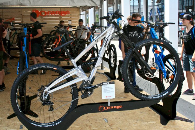 A display of mountain bikes at an outdoor event, featuring a prominent silver bike with a distinctive frame at the forefront. Other bikes in various colors, including blue and orange, are visible in the background. Several people are observing and interacting with the bikes, with a wooden backdrop displaying the brand name "Orange." The setting is lively, showcasing the latest in cycling technology.