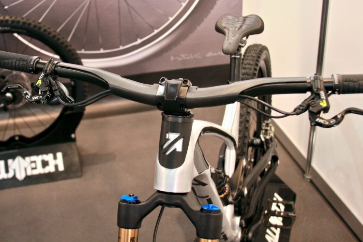 Alt text: Close-up view of a mountain bike's handlebars, including grips and brake levers, with a black and silver frame and forks in the background. A blurred wheel is visible behind, and a logo on the bike's frame is partially shown.