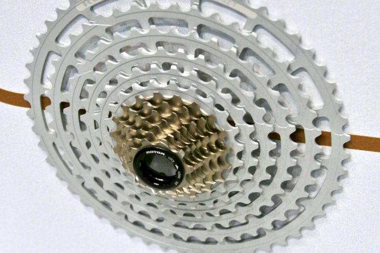 Close-up of a multi-gear bicycle cassette featuring a gold and silver design, labeled "MTB / 10-52". The cassette is mounted on a display board with a white background, highlighting its intricate tooth patterns and central locking mechanism.