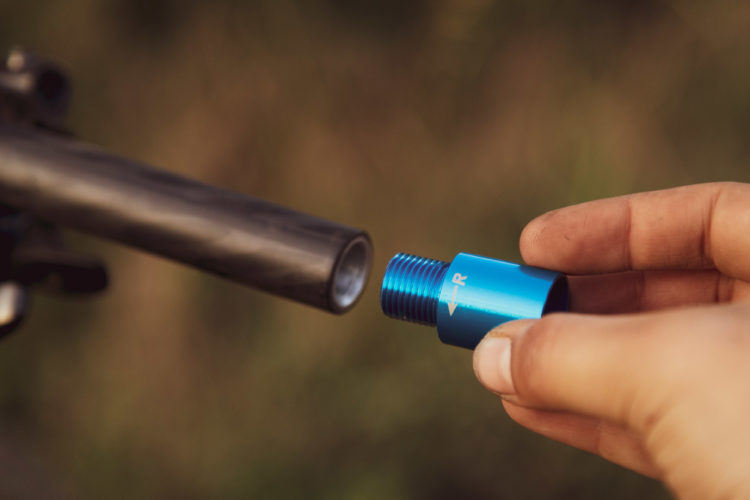 A close-up image of a person's hand holding an aluminum blue threaded accessory, approaching a black firearm's barrel. The background is blurred, suggesting an outdoor setting.