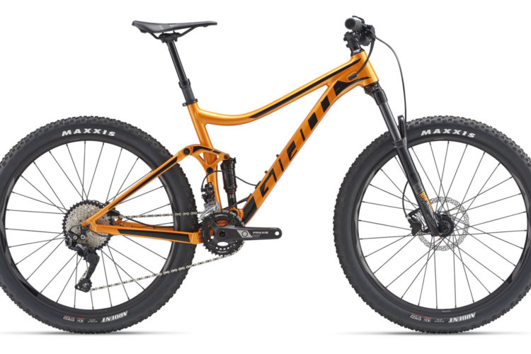 Alt text: A bright orange mountain bike with a sleek design, featuring large black tires labeled "Maxxis" and a modern gear system. The bicycle is positioned against a plain white background.