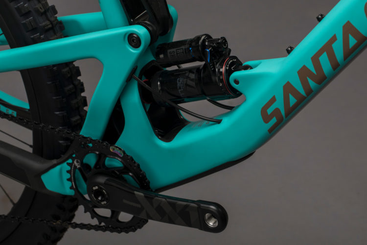 Close-up view of a turquoise bicycle frame featuring the brand logo "SANTA." The image showcases the bike's rear suspension system with two shock absorbers, chainring, and cranks. The background is a neutral gray, emphasizing the bike's design and components.