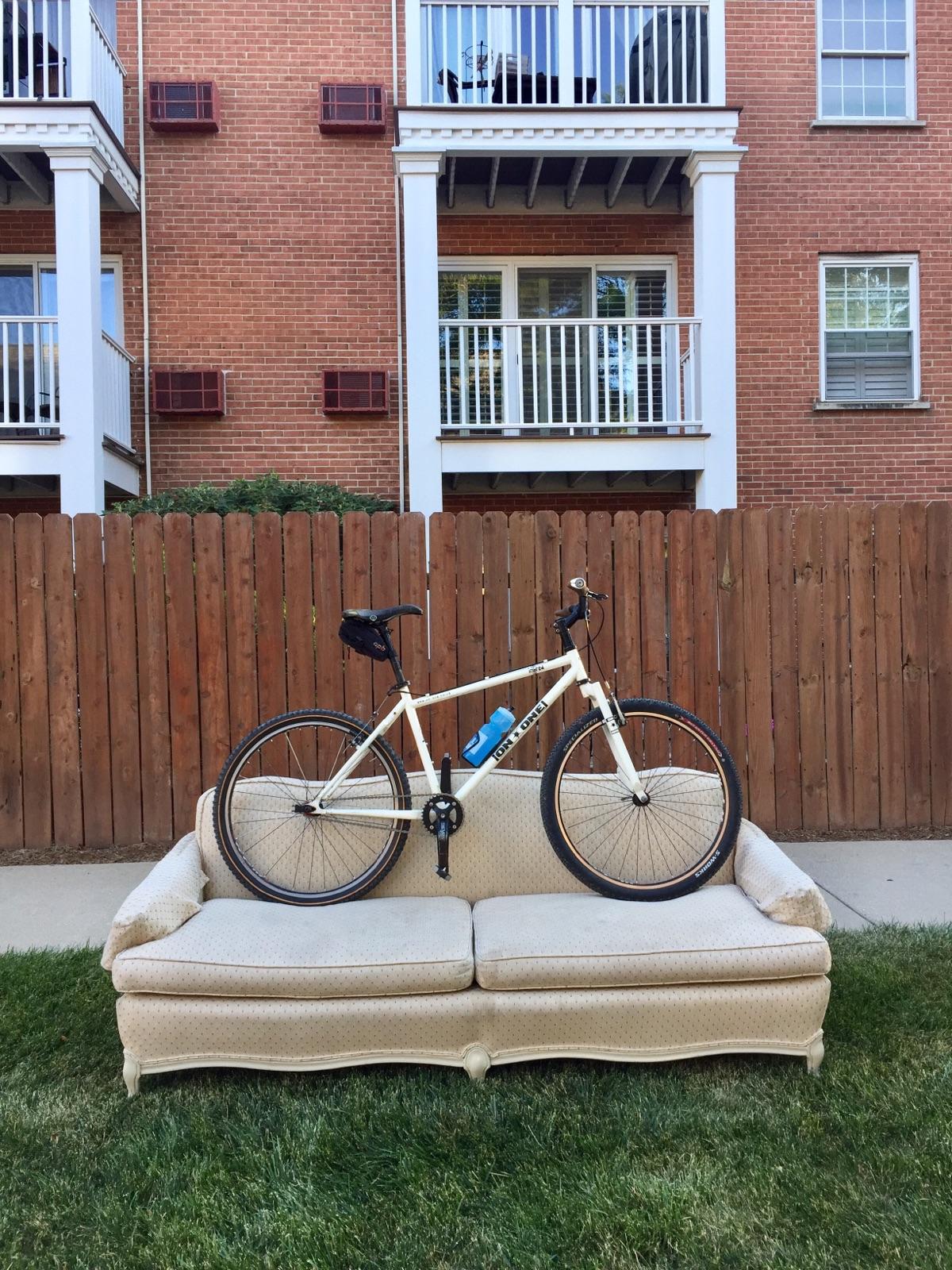 On-One On one inbred: A white bicycle is placed on a beige couch, set against a backdrop of a wooden fence and brick buildings with balconies. The scene displays a mix of outdoor elements, including green grass and decorative features of the apartment complex.