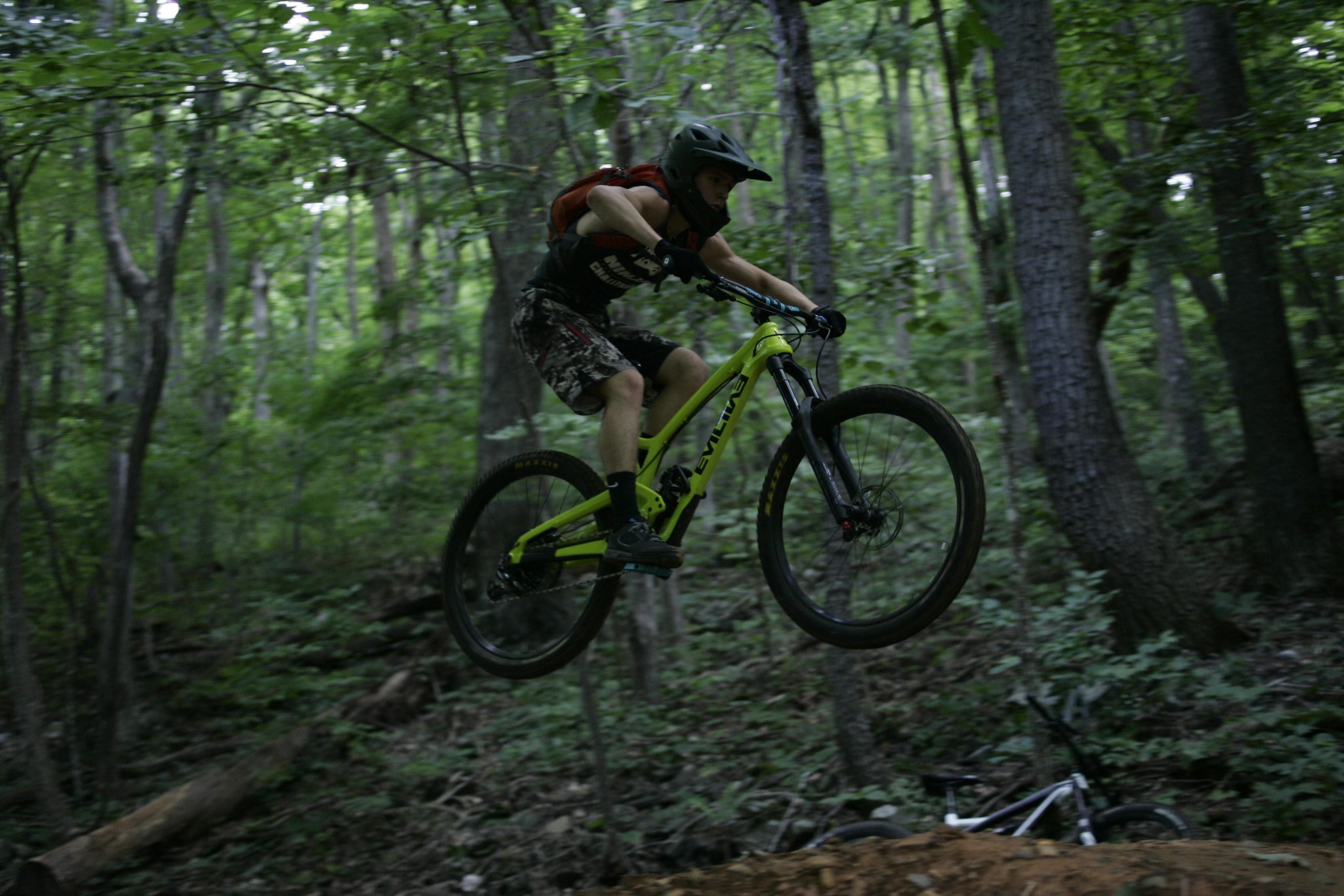 A mountain biker in a helmet performs a jump on a vibrant green bike in a dense forest. The rider is airborne above a dirt path, with trees and underbrush in the background. Another bike is partially visible on the ground nearby. Rocky Knob Park mountain bike trail.