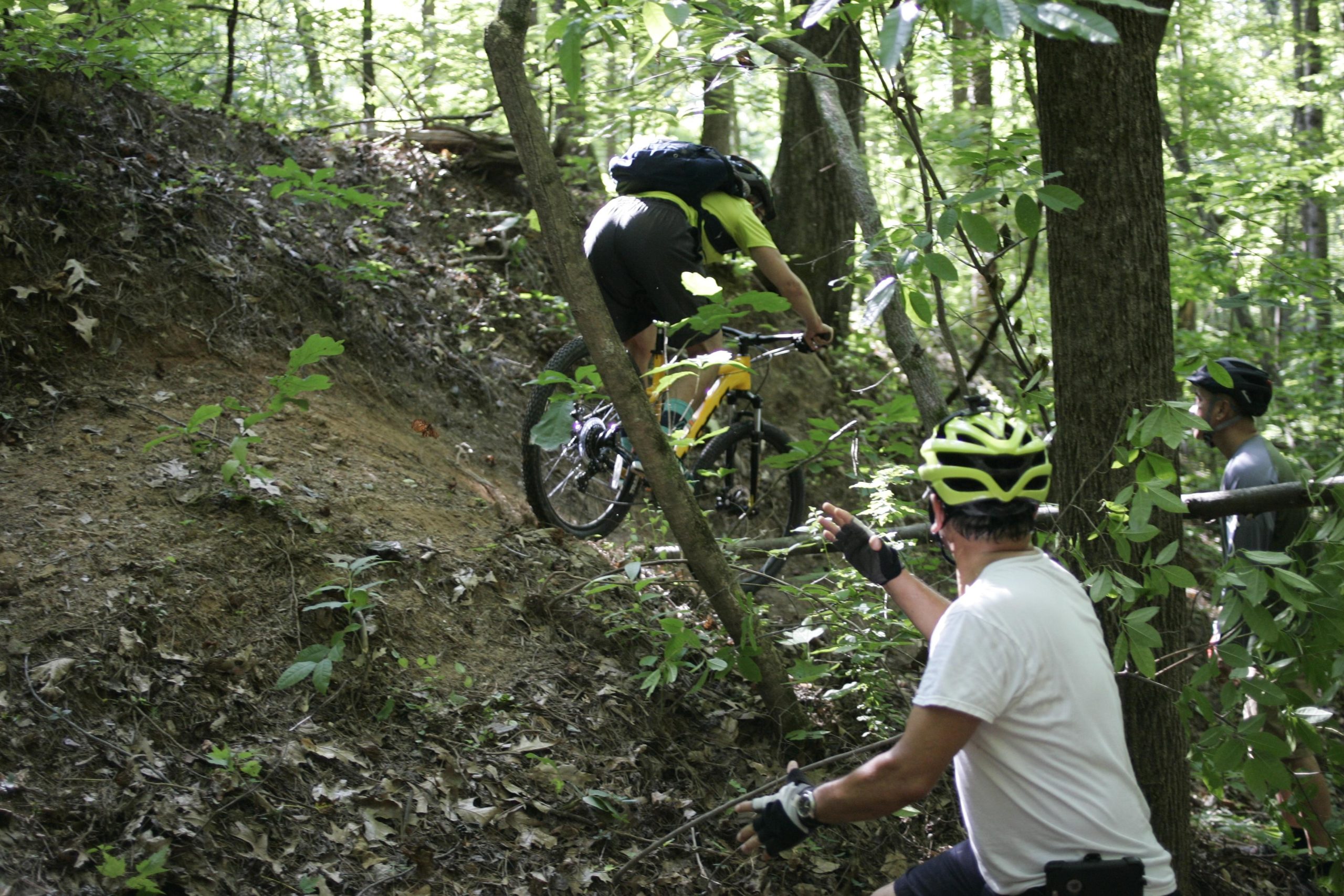 A cyclist navigates a steep, muddy incline in a wooded area while two onlookers offer encouragement. The scene captures the intensity and excitement of mountain biking in nature, surrounded by lush green foliage. Salem Lake mountain bike trail.
