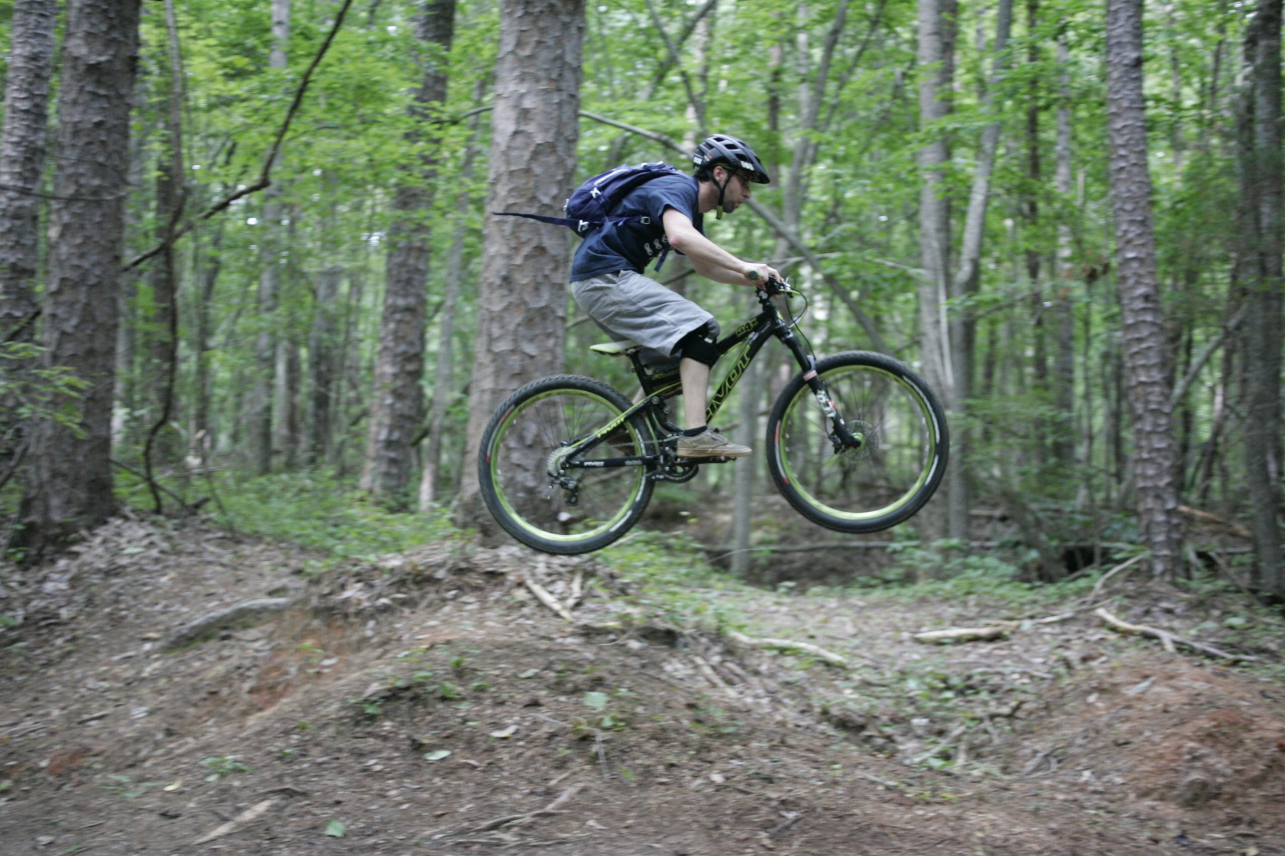 A mountain biker mid-air performing a jump on a trail surrounded by dense green forest. The cyclist wears a helmet, a dark shirt, and shorts, and is riding a black mountain bike with yellow accents. Salem Lake mountain bike trail.