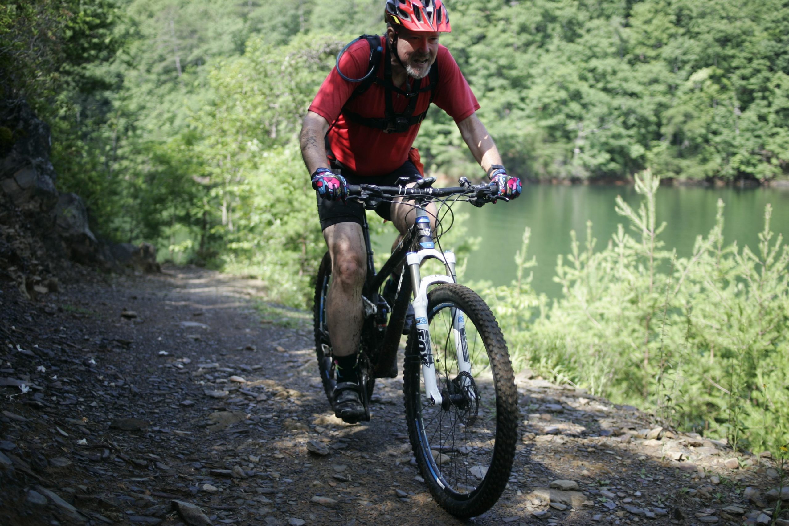 A cyclist in a red shirt and helmet rides a mountain bike along a rocky trail, surrounded by greenery and a serene body of water in the background. The cyclist appears focused and is equipped with protective gear, showcasing an active outdoor lifestyle. Tsali Recreation Area mountain bike trail.