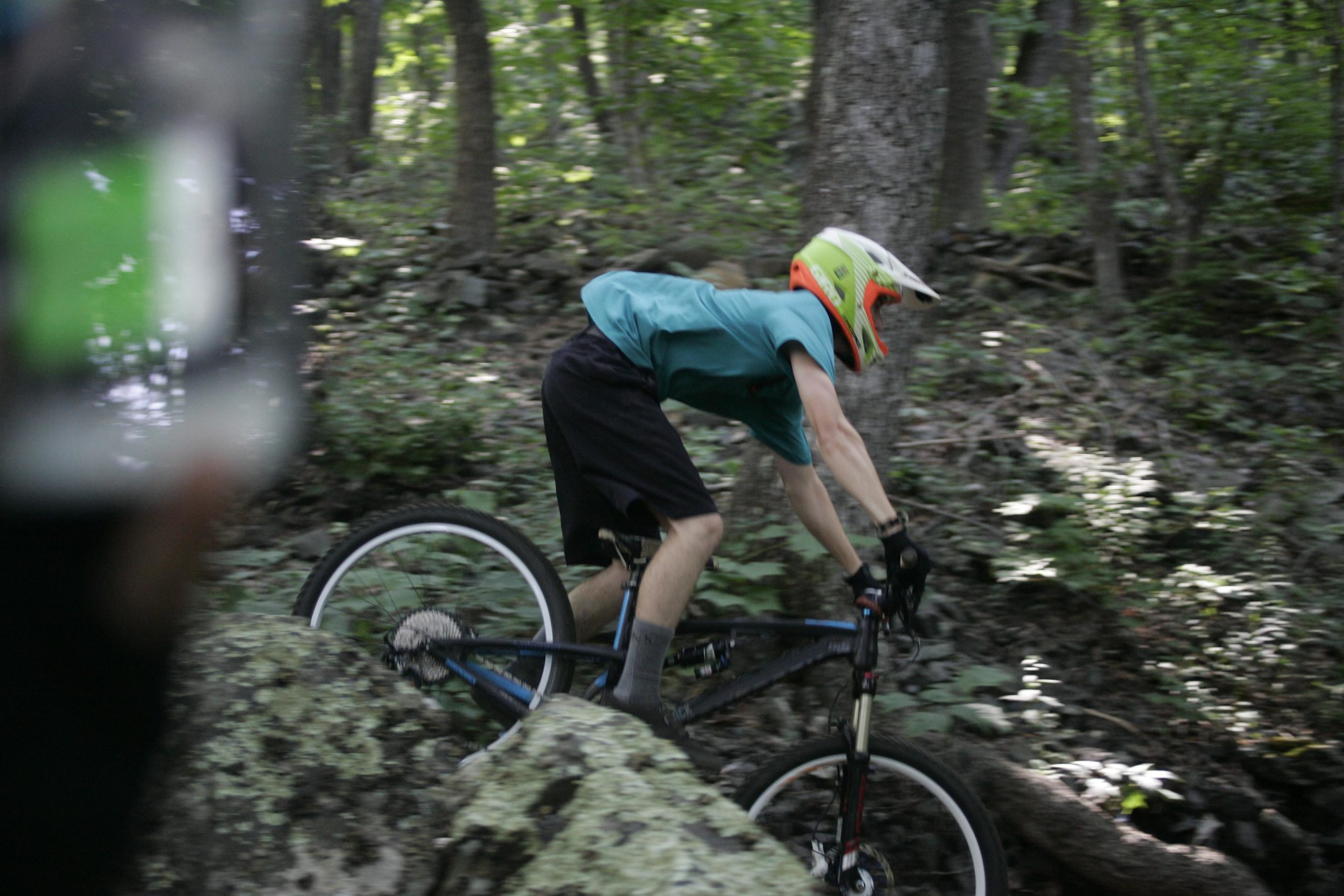 A young cyclist in a bright helmet rides a mountain bike over a rocky terrain in a wooded area, demonstrating balance and skill. The scene is set in a lush, green environment with trees and shadows. Rocky Knob Park mountain bike trail.