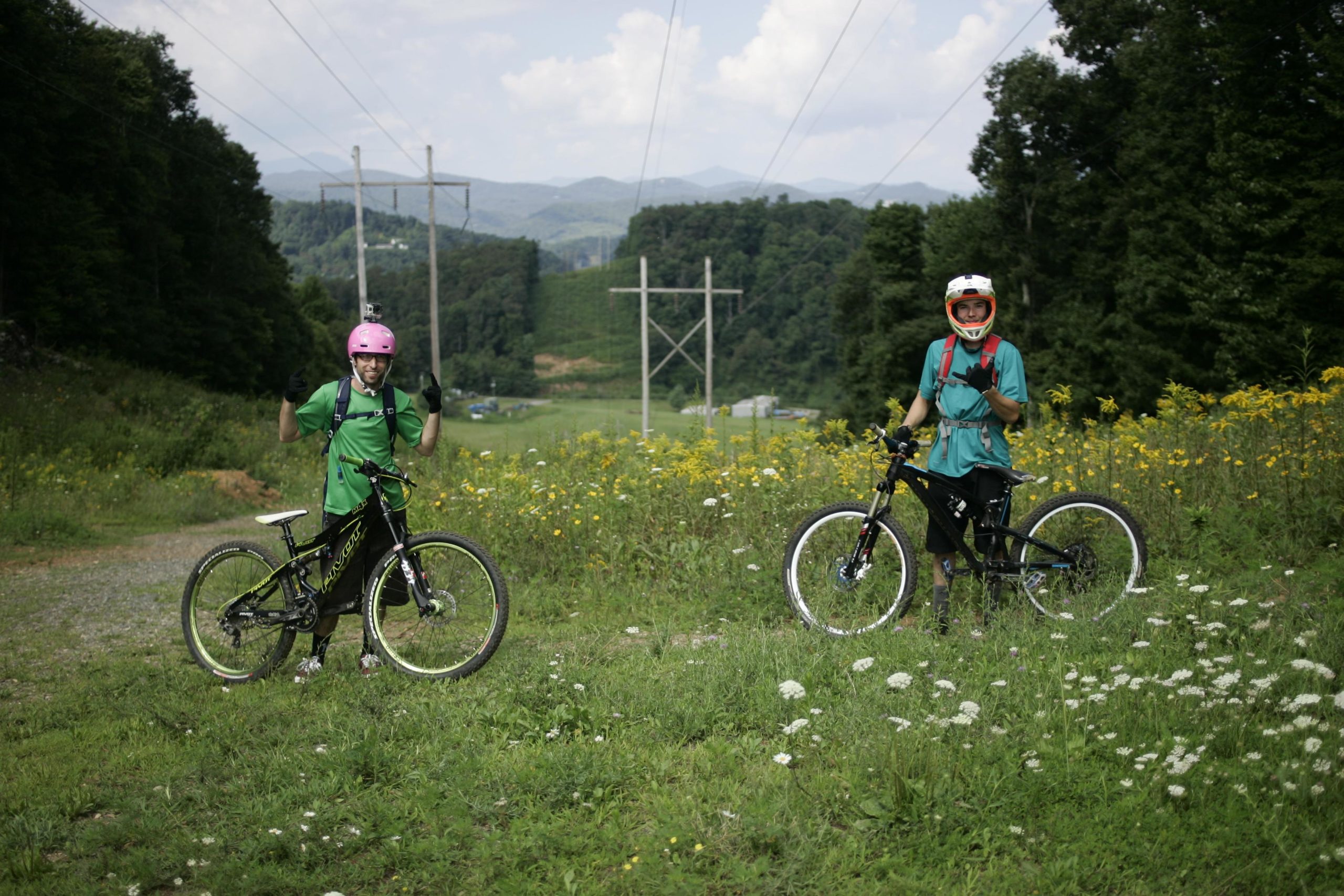 Two mountain bikers stand on a grassy trail surrounded by wildflowers, smiling and giving thumbs up. One rider wears a pink helmet and a green t-shirt, while the other wears a red helmet and a blue jersey. In the background, power lines and rolling hills are visible under a partly cloudy sky. Rocky Knob Park mountain bike trail.