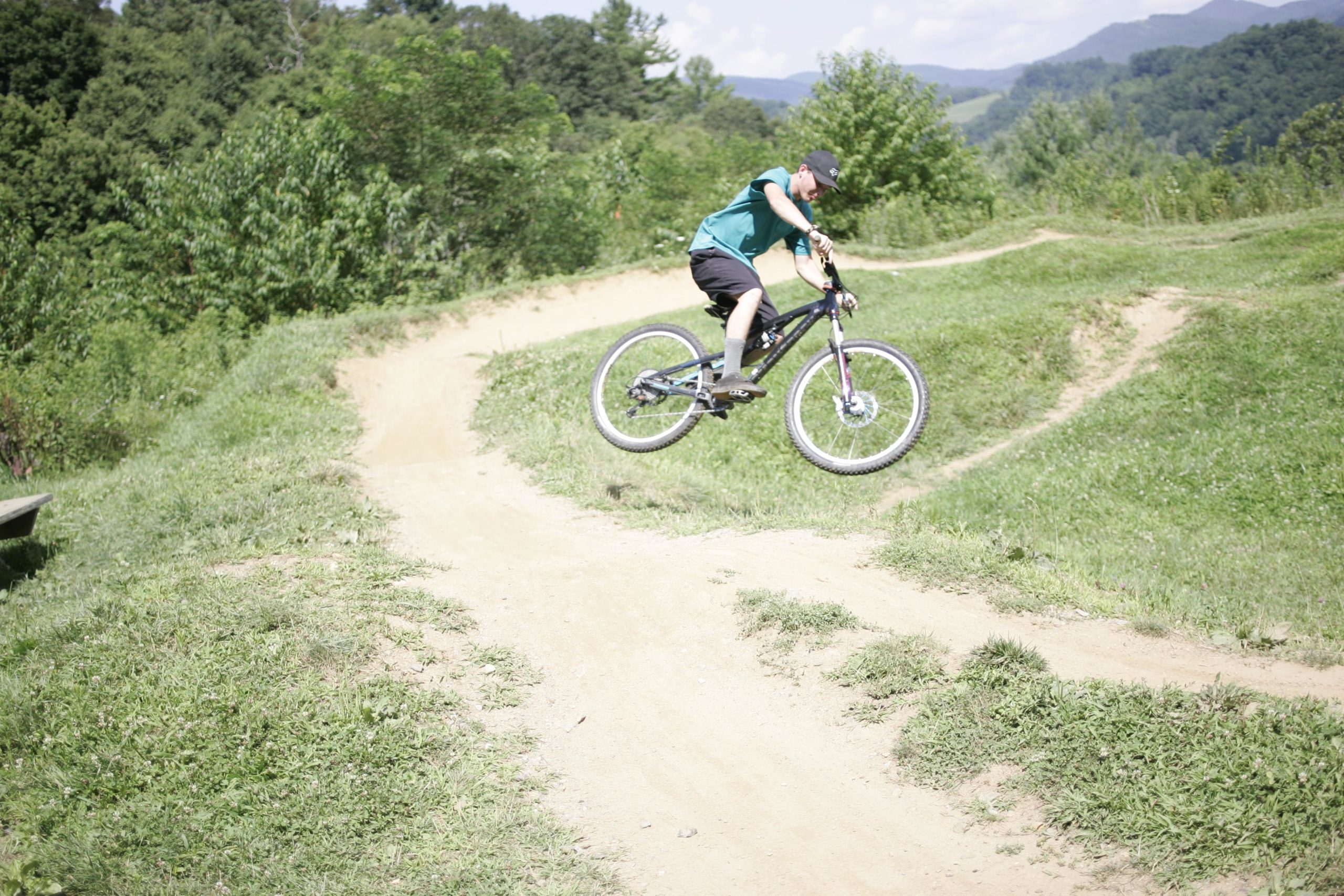 A young rider in a teal shirt and black shorts performs a jump on a mountain bike over a dirt trail in a grassy area, with greenery and mountains in the background. The scene captures the excitement of biking outdoors. Rocky Knob Park mountain bike trail.