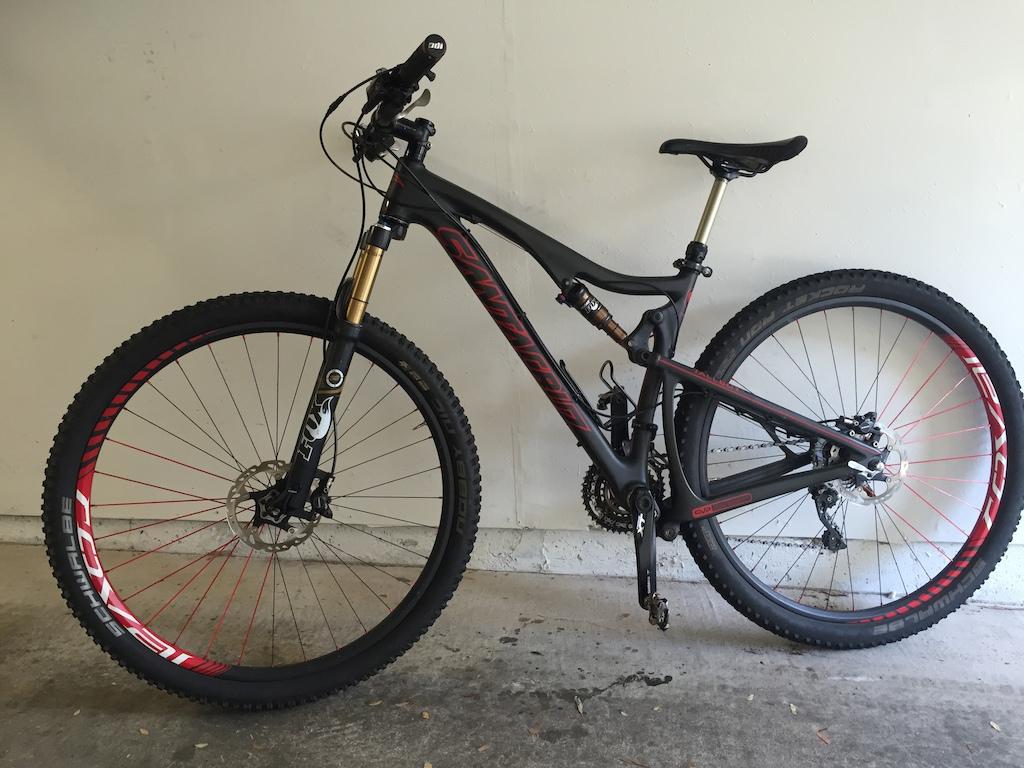 Santa Cruz Tallboy Carbon: Mountain bike with a black and red frame, featuring front suspension and thick, knobby tires. The bike is positioned upright against a plain wall, displaying its detailed components such as the handlebars, gears, and disc brakes.