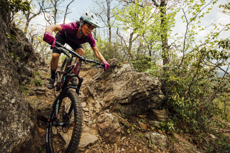 A mountain biker navigating a rocky trail surrounded by greenery, wearing a helmet and vibrant cycling gear, focused on maintaining balance as they tackle an uneven terrain. Trees and foliage are visible in the background under a cloudy sky.