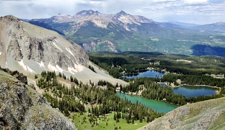 A panoramic view of a mountainous landscape featuring rugged peaks, lush green valleys, and several glistening lakes. The foreground shows rocky terrain with patches of snow, while the background reveals a mix of evergreen forests and expansive mountain ranges under a partially cloudy sky.