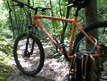 An orange mountain bike parked on a dirt trail surrounded by green trees and foliage, with a black and orange backpack on the ground nearby. Lost Valley mountain bike trail.