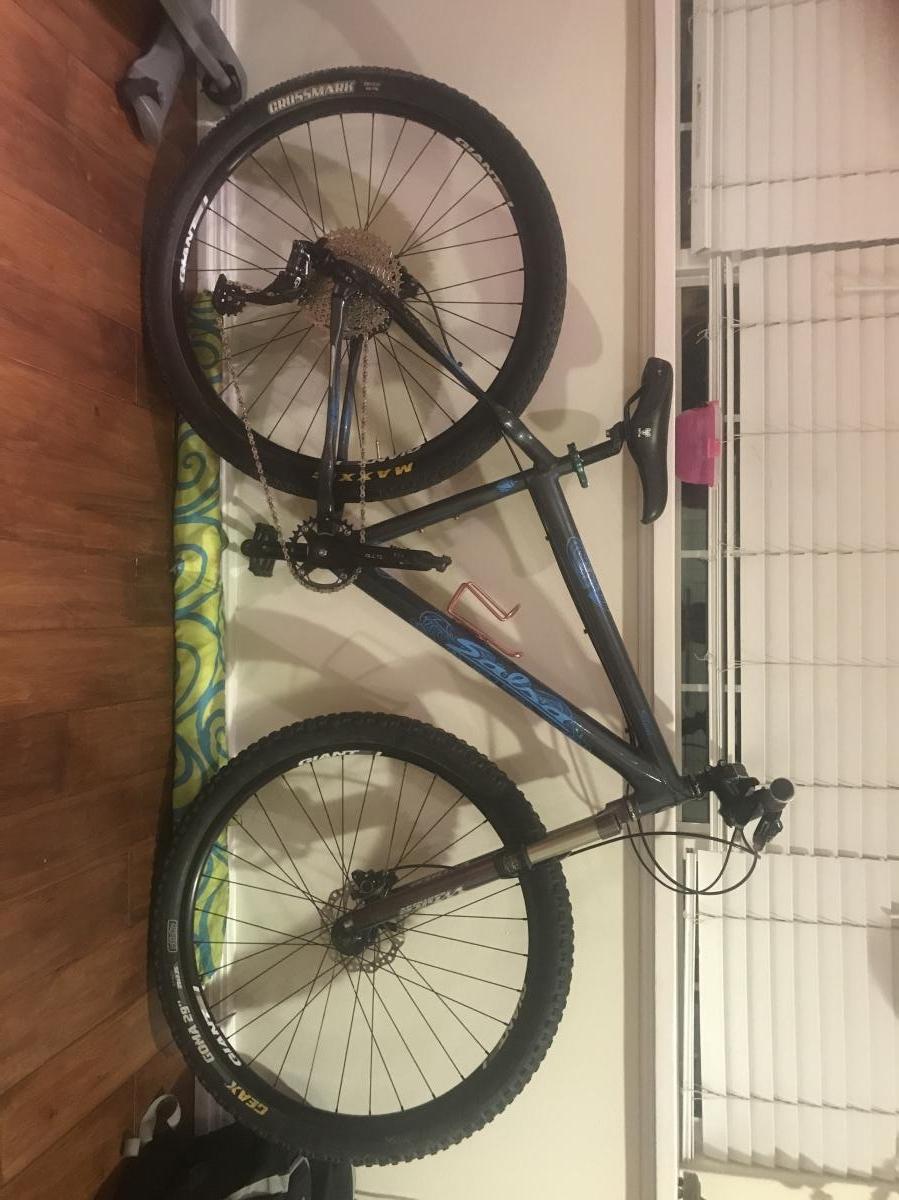 Salsa Selma: A mountain bike leaning against a wall with a wooden floor. The bike features a black frame with blue graphics, wide tires, and a visible gear mechanism. The background includes a partially open window with blinds and a colorful patterned item on the floor.