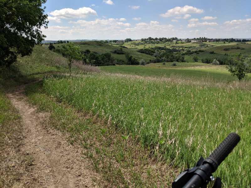 A winding dirt trail leads through a lush green meadow, framed by trees on the left. The landscape features rolling hills and distant greenery under a bright blue sky with scattered white clouds. A bicycle handlebar is visible in the foreground, indicating an outdoor adventure. Sunset Park trail (AKA Eagle Trail) mountain bike trail.