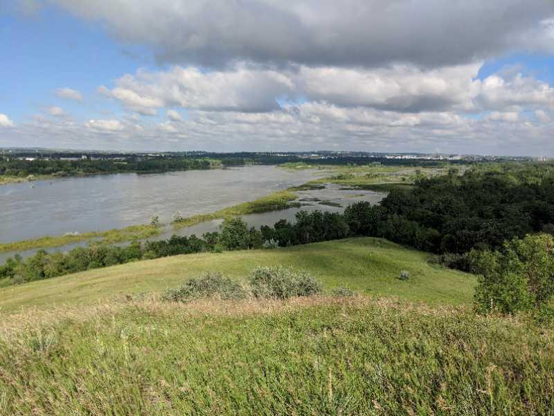 A scenic view of a river winding through lush green hills, with bright blue skies dotted with clouds. The landscape features a mix of trees and grass in the foreground, with the river visible in the middle ground, showcasing a tranquil natural setting. Pioneer Park Main Trail mountain bike trail.