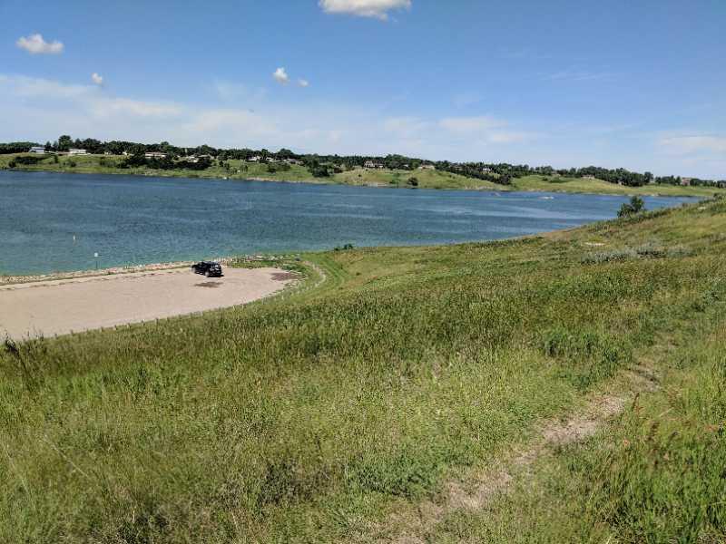 A serene landscape featuring a grassy hillside leading to a calm lake under a clear blue sky, with a few clouds visible. In the foreground, a sandy area is visible near the water, and a vehicle is parked nearby. The lush green hills in the background are dotted with trees and houses. Overlook Trail mountain bike trail.