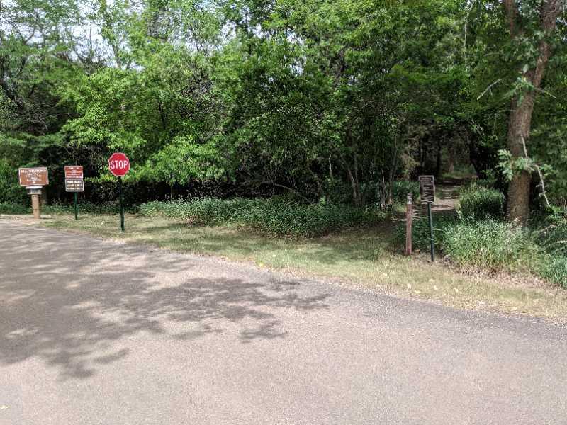A gravel road intersects a lush, green area with trees and bushes. On the left, a red stop sign is prominently displayed, and two brown informational signs can be seen nearby, indicating rules or information about the area. A narrow path leads into the greenery on the right, inviting exploration. Sunlight filters through the leaves, creating dappled shadows on the ground. Richmond Lake Forest Drive mountain bike trail.