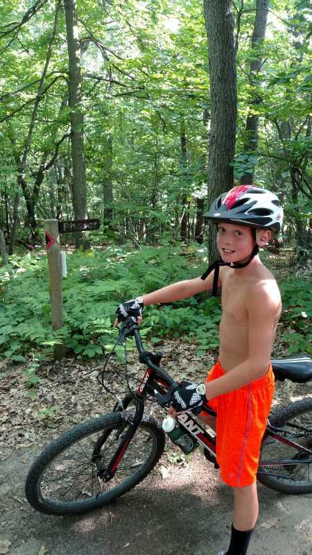 A young boy wearing a black and red helmet and black gloves stands beside his mountain bike on a forest trail. He is shirtless and wearing bright orange shorts, smiling while facing the camera. Behind him, a wooden trail sign and lush green foliage can be seen, indicating a biking path through the trees. Merrell Trail mountain bike trail.