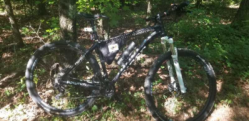 A black mountain bike resting on the ground in a forested area, surrounded by green foliage and trees. The bike has a white front fork and is equipped with a small bag attached to the frame. Sunlight filters through the trees, illuminating the scene. Yellow River mountain bike trail.