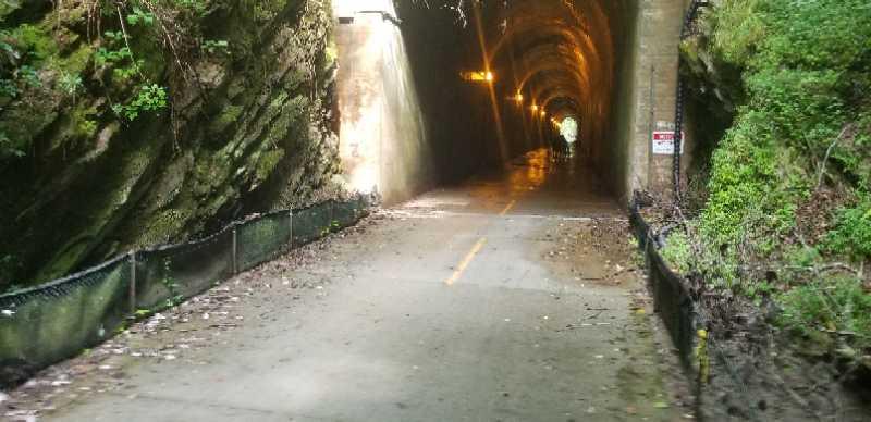 A dimly lit tunnel along a biking and walking path, with rocky walls and greenery on either side. The pavement is visible, showing a bike lane, and there are lights illuminating the tunnel's interior. A sign is partially visible, indicating the area. The scene suggests a natural environment transitioning into an enclosed space. Silver Comet Rail Trail mountain bike trail.