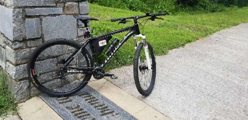 A black mountain bike with a battery pack attached is leaning against a stone structure near a grassy area. A path is visible in the background, with the words "Welcome to..." partly visible on a stone slab near the bike. Silver Comet Rail Trail mountain bike trail.