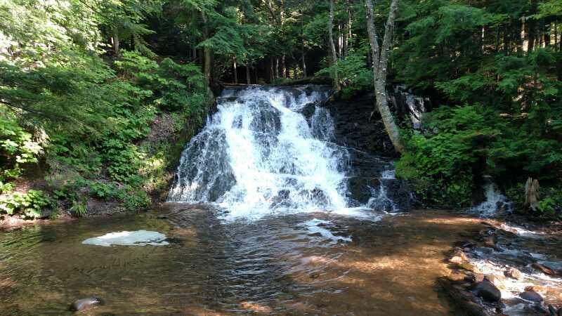 Alt text: A serene water cascade flows over rocks into a clear pool, surrounded by lush green foliage and trees under dappled sunlight. Noquemanon Trails Network: South Marquette Trails mountain bike trail.