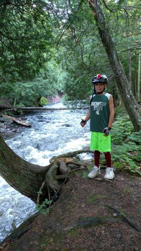 A child wearing a helmet stands on a bank by a flowing river, surrounded by lush green trees. The child is dressed in a Michigan State tank top and bright green shorts, with a cheerful expression. The scene captures a moment of outdoor adventure in a natural setting. Noquemanon Trails Network: South Marquette Trails mountain bike trail.
