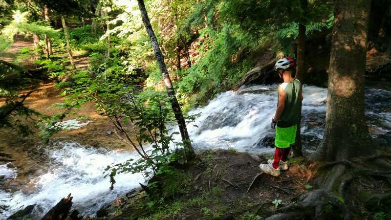 A child wearing a helmet and colorful clothing stands at the edge of a flowing river in a forested area. The scene features lush green foliage, with sunlight filtering through the trees, creating a peaceful outdoor atmosphere. Noquemanon Trails Network: South Marquette Trails mountain bike trail.
