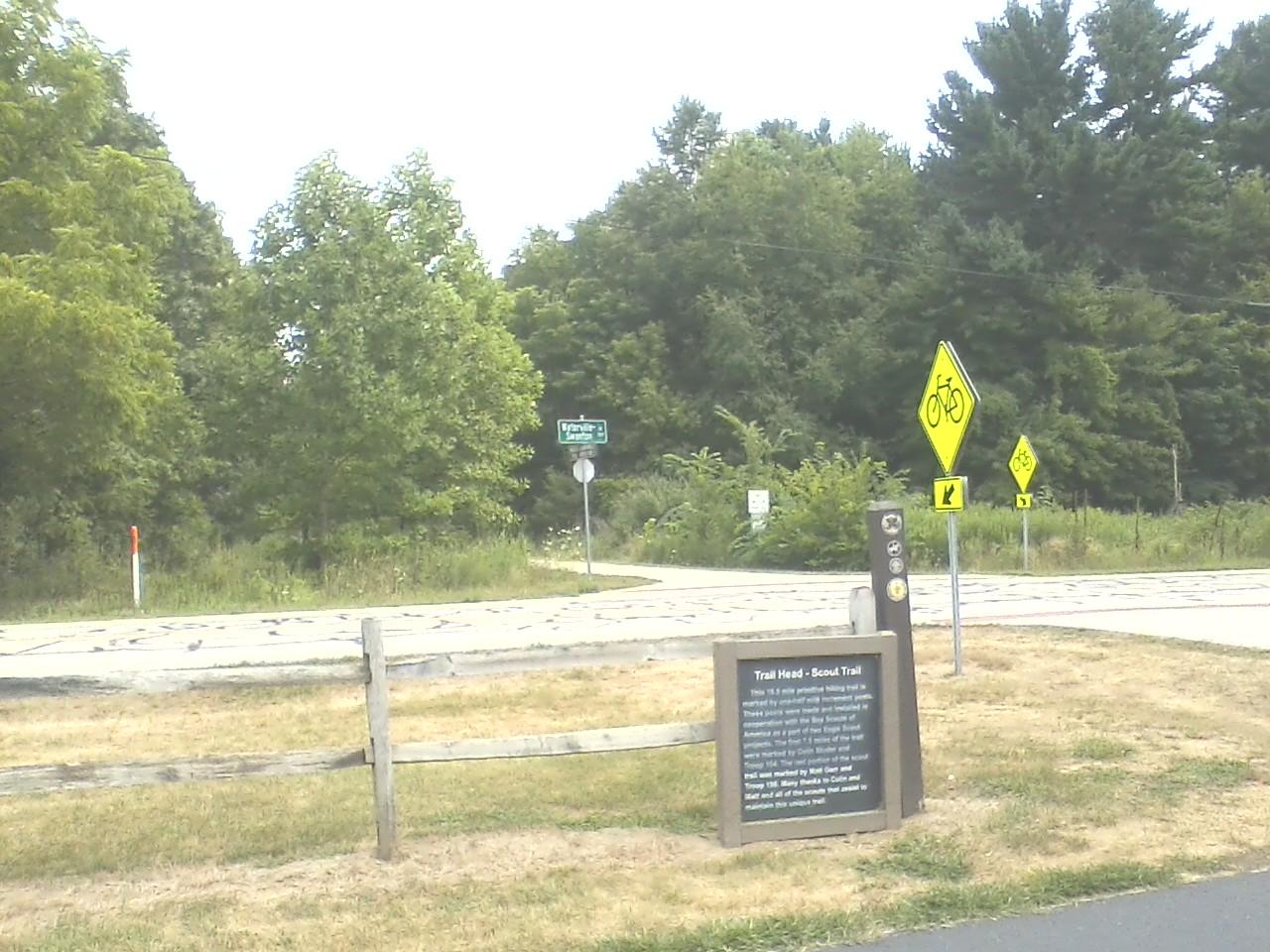 Trailhead sign for Scout Trail located at an intersection, surrounded by greenery. The sign provides information about the trail, including its length and features. Bicycle warning signs are visible, indicating a bike-friendly area. Oak Openings -- Beach Ridge Singletrack Trail mountain bike trail.